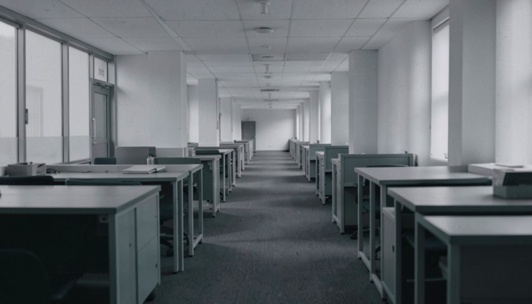Wide duotone photograph in muted slate-purple and teal of an empty office corridor with rows of desks fading into the distance