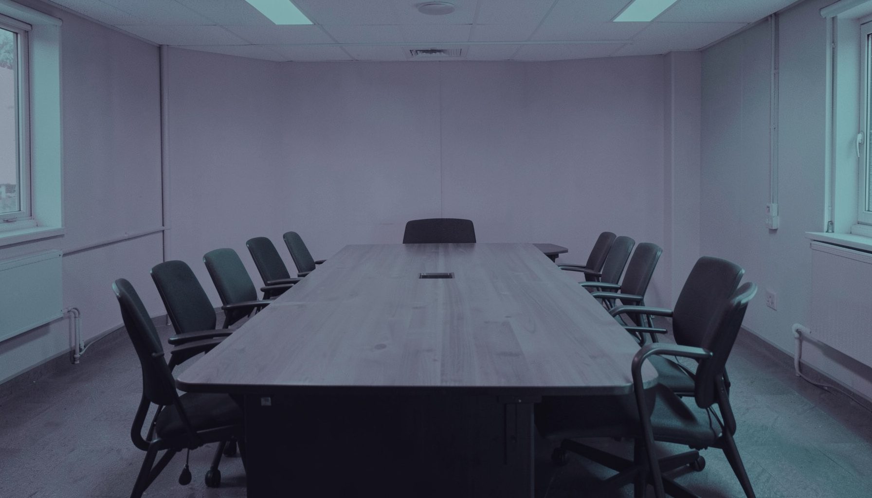 Duotone slate-purple and teal photograph of a meeting room with empty chairs around a long wooden table, calm institutional setting