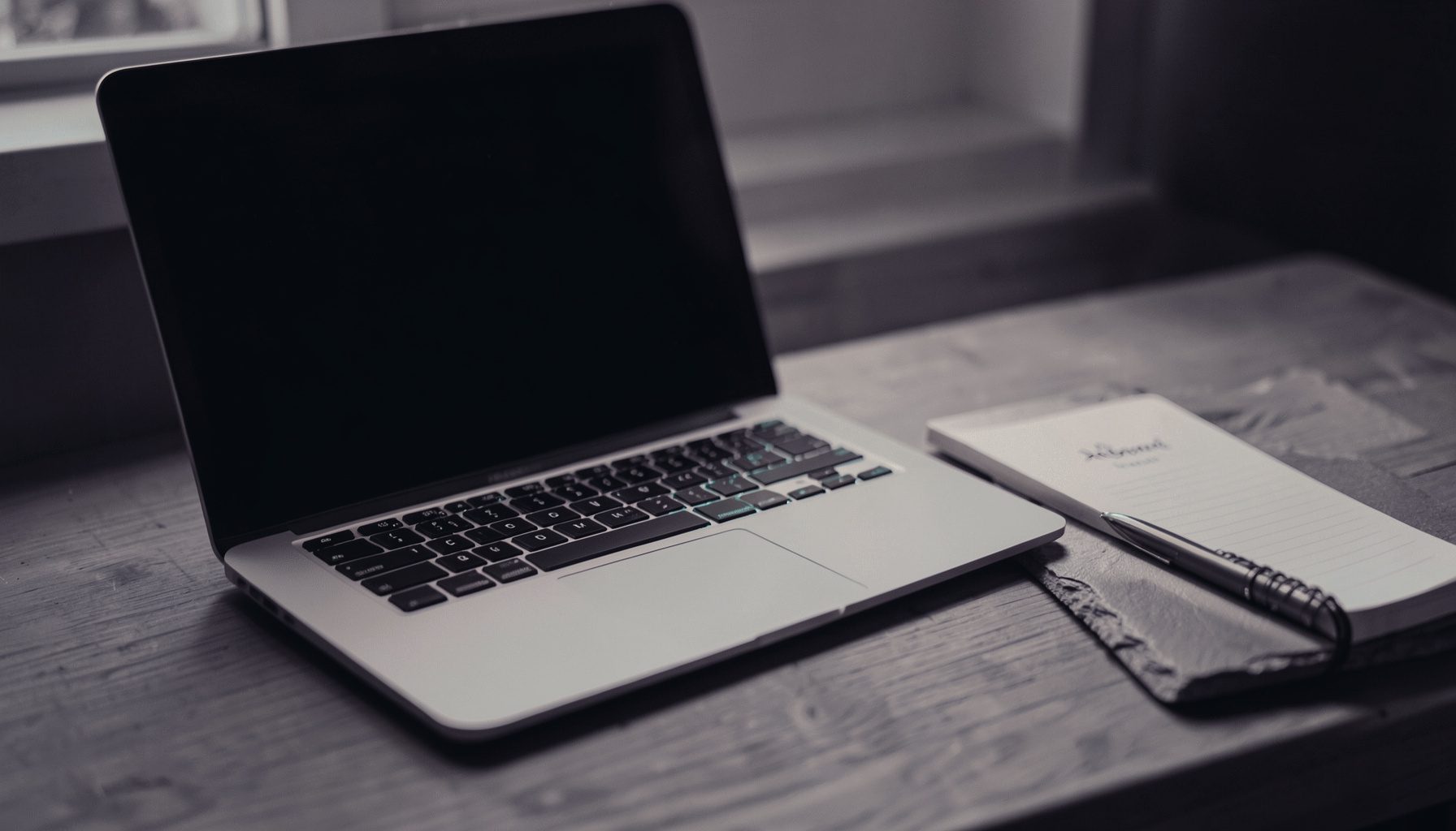 Duotone slate-purple and teal photograph of a laptop open on a wooden desk beside a notebook, warm muted light, calm professional workspace