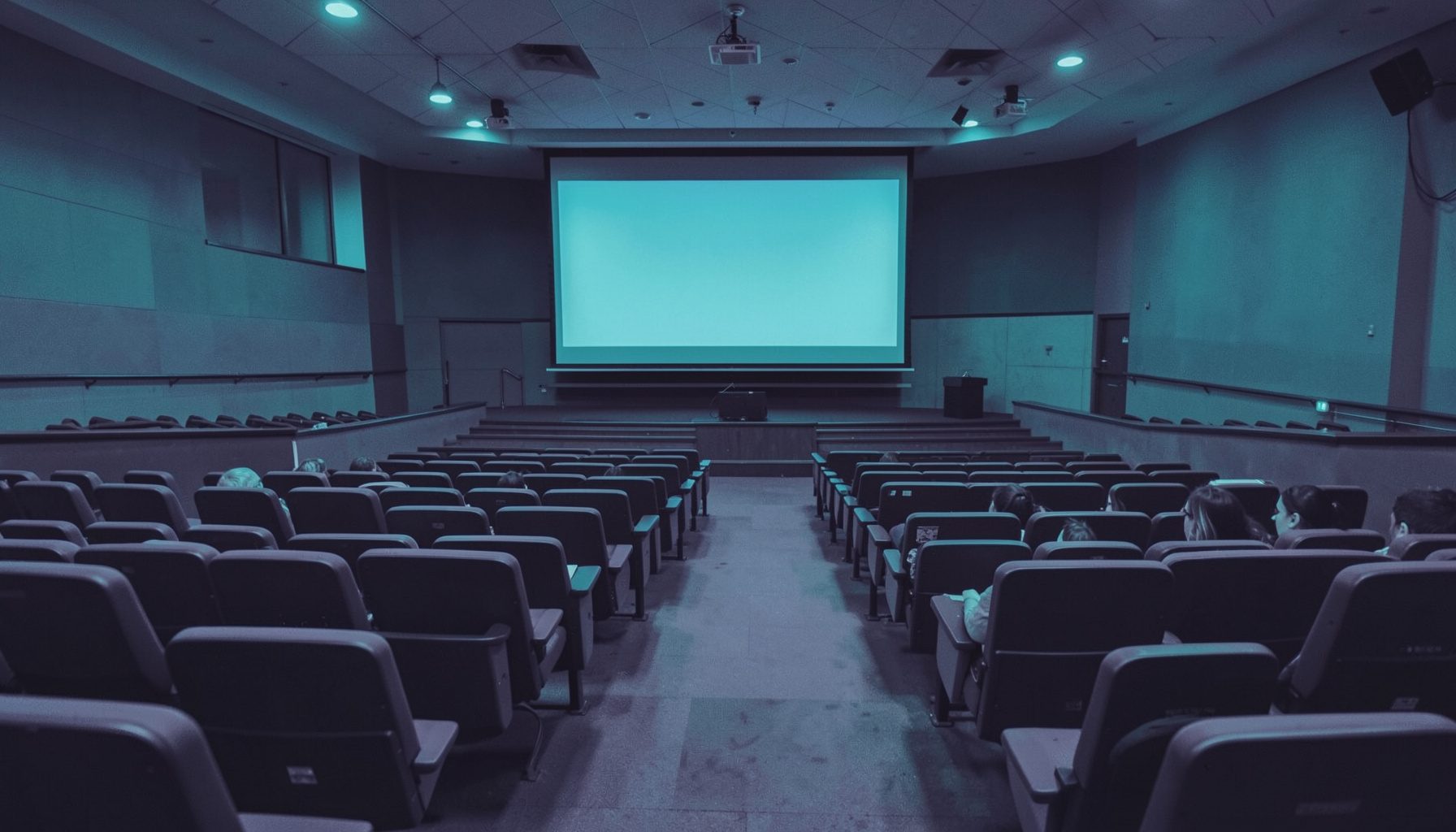 Duotone slate-purple and teal photograph of an empty lecture hall with tiered seating and a presentation screen at the front, soft institutional light