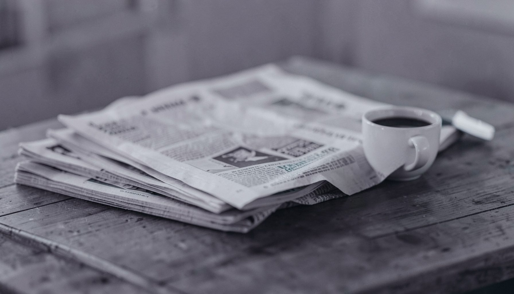 Duotone slate-purple and teal photograph of a stack of newspapers and a coffee cup on a wooden table, soft diffused daylight
